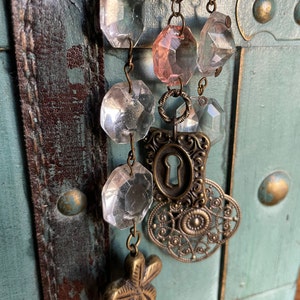 Puede incluir: Un collar de inspiraci&oacute;n vintage con cuentas de vidrio transparentes y rosadas, un dije de ojo de cerradura y un dije de metal decorativo. El collar est&aacute; colgado en una superficie de madera desgastada.