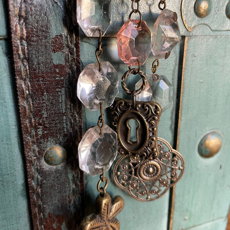 Puede incluir: Un collar de inspiraci&oacute;n vintage con cuentas de vidrio transparentes y rosadas, un dije de ojo de cerradura y un dije de metal decorativo. El collar est&aacute; colgado en una superficie de madera desgastada.