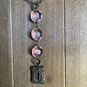 Puede incluir: Un collar de cristal rosa con un dije de ojo de cerradura. El collar tiene una cadena con cuatro cuentas de cristal rosa y un dije de ojo de cerradura en la parte inferior.