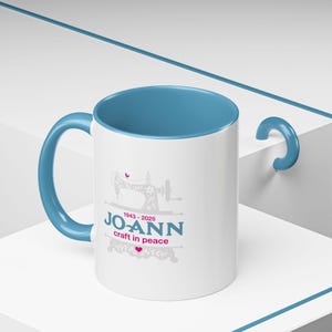 May include: White ceramic mug with a blue interior and handle. The mug features a sewing machine graphic and the text "JO-ANN craft in peace" in teal and pink, with the years 1943-2025.
