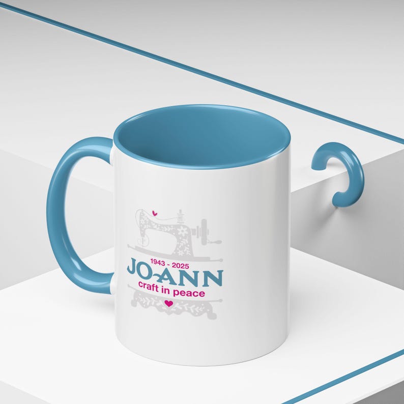 May include: White ceramic mug with a blue interior and handle. The mug features a sewing machine graphic and the text "JO-ANN craft in peace" in teal and pink, with the years 1943-2025.