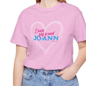 Könnte beinhalten: Hellrosa T-Shirt mit herzförmigem Aufdruck. Der Aufdruck enthält den Text "I miss my friend JOANN" in Rosa und Blau, mit den Jahren "1943-2023" darunter. Das T-Shirt wird mit einer blauen Jeans getragen.