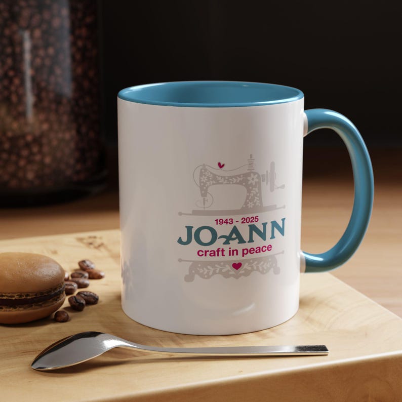 May include: White ceramic mug with a teal interior and handle. The mug features a sewing machine graphic and the text "JO-ANN craft in peace" with the years 1943-2025. A spoon and a macaron are in the foreground.