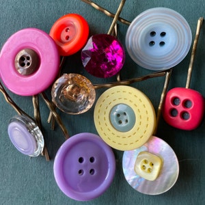 K&ouml;nnte beinhalten: Sortiment aus bunten Kn&ouml;pfen und Haarnadeln. Die Kn&ouml;pfe sind in verschiedenen Formen und Gr&ouml;&szlig;en, darunter rund, quadratisch und oval. Die Farben umfassen Pink, Rot, Blau, Gelb und Lila. Die Haarnadeln sind aus goldenem Metall.