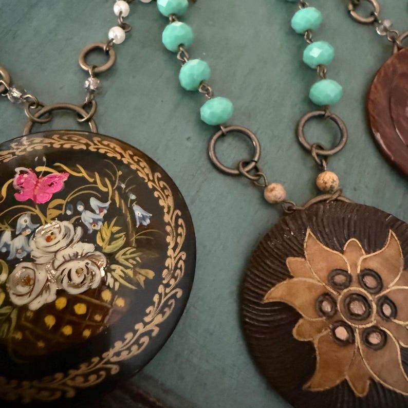 May include: Two necklaces with pendants. One pendant is black with gold accents and a painted floral design. The other pendant is brown with a carved flower design.