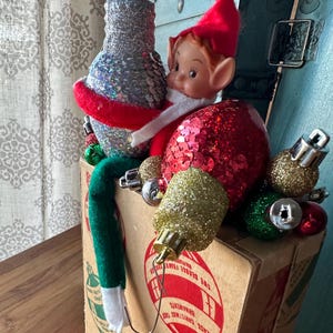 May include: A vintage Christmas ornament box overflowing with holiday decorations. The box contains a silver sequined vase, a red sequined ornament, and various gold and green ornaments. A small elf doll with a red hat and green legs is nestled among the decorations.
