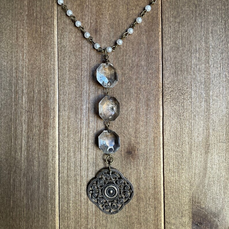 Puede incluir: Un collar largo de cadena plateada con cuentas blancas y un colgante plateado. El colgante presenta tres oct&oacute;gonos de vidrio transparente y un dise&ntilde;o de filigrana decorativo.