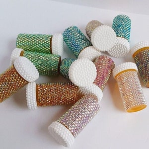 May include: A collection of small, clear plastic pill bottles decorated with iridescent rhinestones. The bottles are arranged in a random pattern on a white surface.