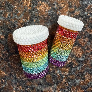 May include: Two decorative pill bottles encrusted with colorful rhinestones in a rainbow gradient. Each bottle has a white, textured, pill-shaped cap. The bottles are set against a dark, speckled background.