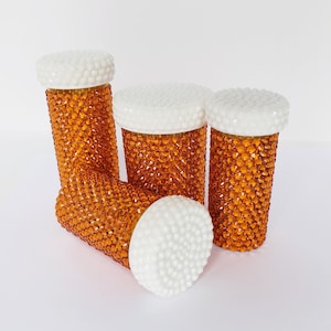 May include: Four amber glass jars with white lids, each covered in orange rhinestones. The jars are arranged in a group, with the lids facing up.