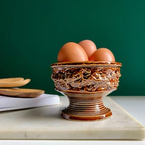 May include: A brown ceramic egg cup with a textured design, containing three brown eggs. The egg cup has a pedestal base and a decorative rim. The background is a dark green colour.