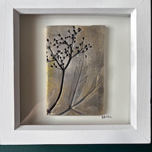 May include: Framed botanical art. The artwork features a textured panel with a pressed plant design. A dark, branching stem with small buds and a leaf impression are set against a muted gold and grey background. The frame is white.
