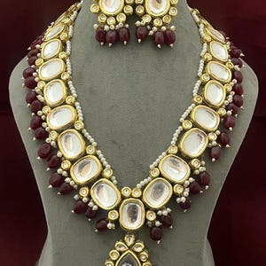 May include: A gold necklace with a teardrop pendant and red beads. The necklace is made of small, round, clear stones set in gold. The pendant is also made of clear stones and has a gold frame. The necklace is paired with matching earrings.