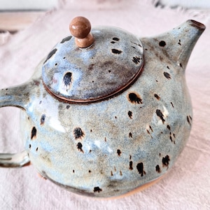 Könnte beinhalten: Eine Keramik-Teekanne mit einer gesprenkelten blau-grauen Glasur. Die Teekanne hat einen runden Körper, einen gebogenen Griff und einen Ausguss. Der Deckel ist mit einem kleinen, braunen Knauf versehen. Schwarze Sprenkel bedecken die Oberfläche.
