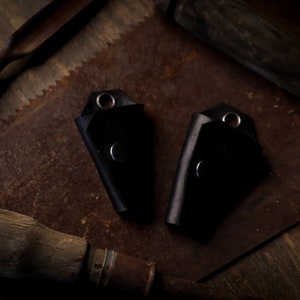 May include: Two black leather key holders with silver rivets and eyelets. The key holders are displayed on a rustic, weathered surface with vintage tools in the background. The key holders are a dark, solid color.