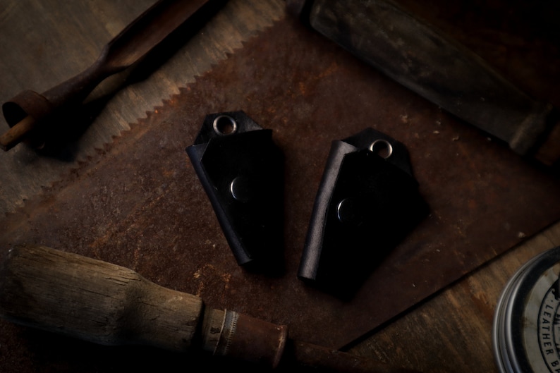 May include: Two black leather key holders with silver rivets and eyelets. The key holders are displayed on a rustic, weathered surface with vintage tools in the background. The key holders are a dark, solid color.