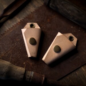 May include: Two light brown leather keychains with brass rivets and eyelets. The keychains are folded into a triangular shape. The background includes vintage tools like a saw and a wooden handle.