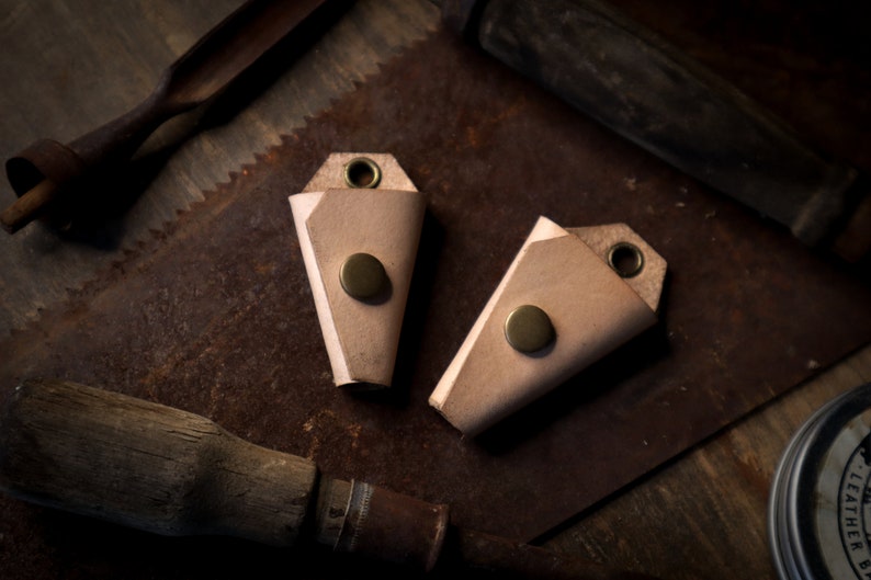 May include: Two light brown leather keychains with brass rivets and eyelets. The keychains are folded into a triangular shape. The background includes vintage tools like a saw and a wooden handle.