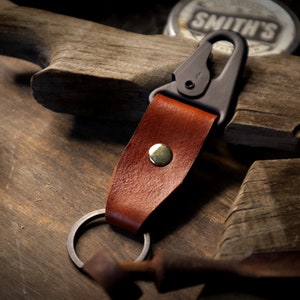 May include: A brown leather key chain with a metal ring and a black metal clip. The keychain has a brass rivet.