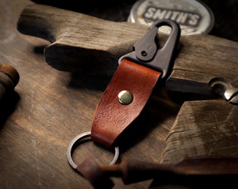 May include: A brown leather key chain with a metal ring and a black metal clip. The keychain has a brass rivet.