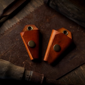 May include: Two brown leather key holders with brass snap closures and eyelets. The key holders are displayed on a rustic, textured surface with vintage tools in the background, creating a handcrafted aesthetic.