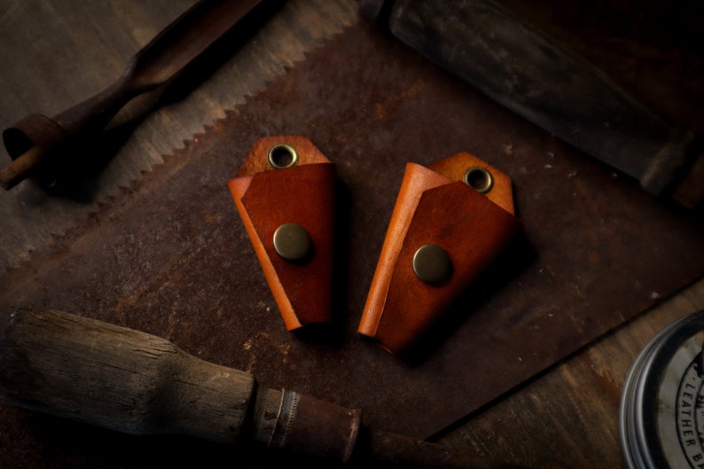 May include: Two brown leather key holders with brass snap closures and eyelets. The key holders are displayed on a rustic, textured surface with vintage tools in the background, creating a handcrafted aesthetic.