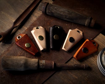 May include: A collection of handmade leather keychains in various colors, including brown, tan, and black, each with a metal rivet. The keychains are arranged with woodworking tools on a rustic, metal surface.