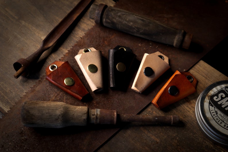 May include: A collection of handmade leather keychains in various colors, including brown, tan, and black, each with a metal rivet. The keychains are arranged with woodworking tools on a rustic, metal surface.