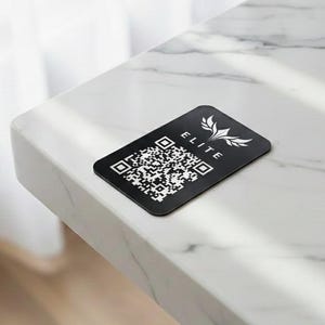 May include: A black rectangular card with a QR code and the word "ELITE" in white lettering. The card has a white floral design and is placed on a white marble surface.