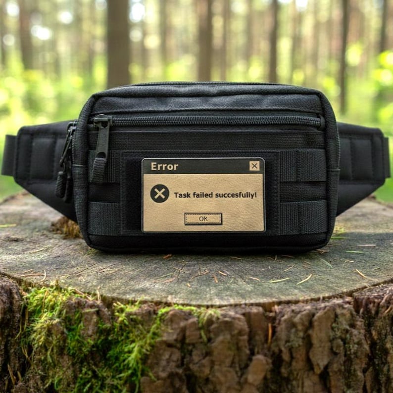 A laser-engraved tan faux leather morale patch featuring the 'Task failed successfully' Windows error meme, attached to a black tactical waist bag sitting on a tree stump in a forest setting.