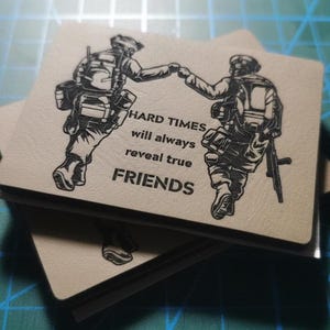 Puede incluir: Una pila de parches cuadrados de color beige con una ilustración en negro de dos soldados chocando los puños. El texto dice "HARD TIMES will always reveal true FRIENDS."