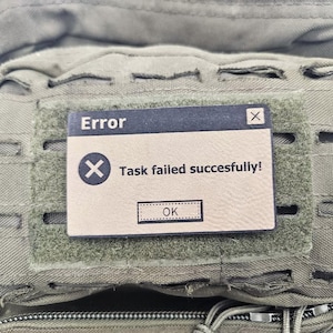 Laser-engraved tan faux leather morale patch with the 'Task failed successfully' Windows error meme, displayed on an olive drab green tactical bag with laser-cut MOLLE webbing