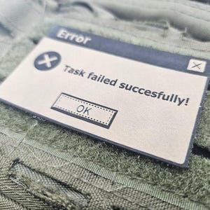 Close-up angled shot of a tan faux leather 'Task failed successfully' morale patch, showing the fine detail of the laser engraving and the clean edges on an olive green tactical surface.