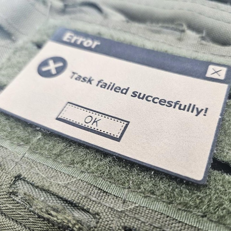 Close-up angled shot of a tan faux leather 'Task failed successfully' morale patch, showing the fine detail of the laser engraving and the clean edges on an olive green tactical surface.
