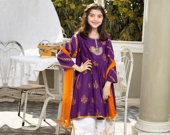 Puede incluir: Una niña posa con un vibrante conjunto bordado en morado y dorado. El conjunto incluye una túnica de manga larga, pantalones blancos con diseños de palmeras doradas y una bufanda naranja. El atuendo es adecuado para ocasiones especiales.
