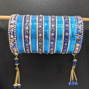 May include: A set of blue bangles with gold and rhinestone accents. The bangles are strung together on a wooden dowel. Each end of the bangles has a gold tassel with blue beads.