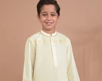 Puede incluir: Un niño con una túnica amarilla clara con bordados en el cuello y los puños, combinada con pantalones blancos y sandalias negras. El conjunto es de estilo tradicional.