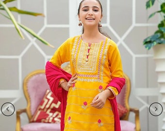 May include: A young person wearing a vibrant yellow salwar kameez ensemble. The outfit features intricate embroidery and pink floral accents. A bright pink scarf is draped over the shoulders. The outfit is paired with brown sandals.