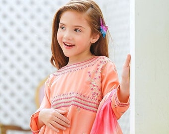Puede incluir: Un vestido color melocotón con detalles bordados y un dupatta a juego. El vestido presenta una silueta acampanada y se combina con pantalones rosas. Una pinza para el pelo con forma de mariposa añade un toque juguetón al conjunto.