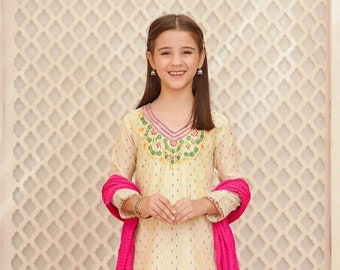 Puede incluir: Una persona joven con un salwar kameez color crema con cuello en V, bordado con motivos florales coloridos. El conjunto incluye una dupatta rosa brillante y pantalones de pernera ancha a juego. El fondo presenta una pared estampada.
