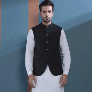 May include: A black waistcoat with a mandarin collar over a white long-sleeve shirt and white pants. The outfit is a traditional South Asian style. The background is a blue wall with a brown stripe.