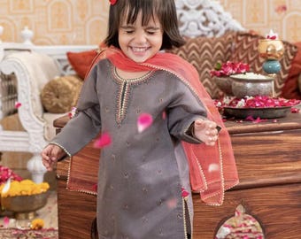 Puede incluir: Una niña pequeña con un atuendo tradicional gris con detalles dorados, que incluye una parte superior de manga larga y una falda estampada. También lleva un lazo rojo y una bufanda naranja transparente. El atuendo está adornado con ribetes dorados.