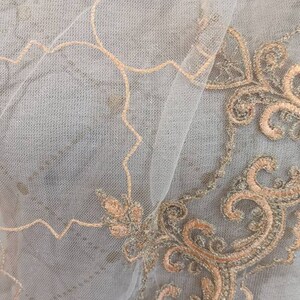 May include: Close-up of a sheer fabric with intricate embroidery. The fabric is a light gray color, with delicate peach-colored embroidery in floral and scroll patterns. The design is detailed and elegant.