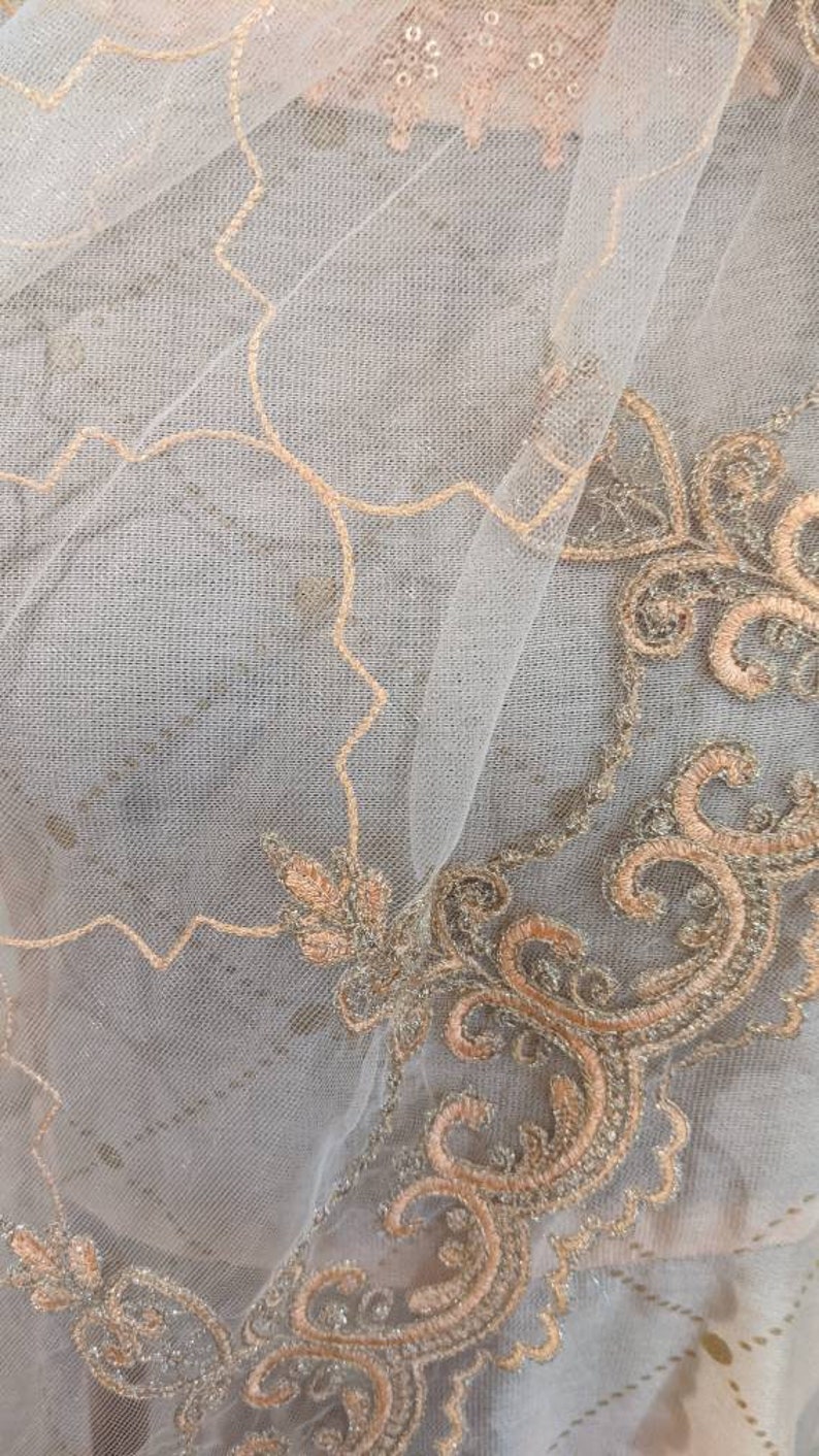 May include: Close-up of a sheer fabric with intricate embroidery. The fabric is a light gray color, with delicate peach-colored embroidery in floral and scroll patterns. The design is detailed and elegant.