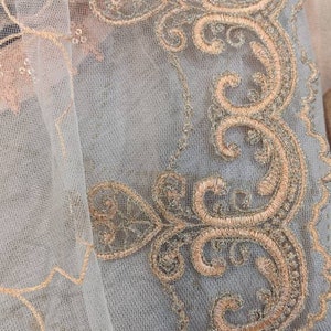 May include: Close-up of a delicate, sheer fabric with intricate embroidery. The design features swirling patterns and floral motifs in shades of gold and peach, creating a luxurious and ornate appearance. The fabric has a soft, textured feel.