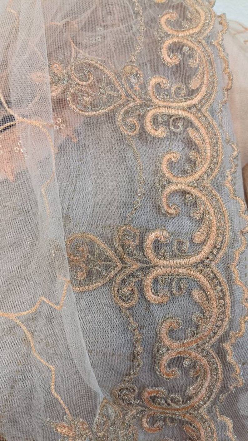 May include: Close-up of a delicate, sheer fabric with intricate embroidery. The design features swirling patterns and floral motifs in shades of gold and peach, creating a luxurious and ornate appearance. The fabric has a soft, textured feel.