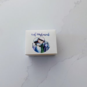 May include: A small white gift box with the words "Eid Mubarak" in blue script. The box features a watercolor illustration of two figures embracing, with a mosque in the background. The box is on a white marble surface.