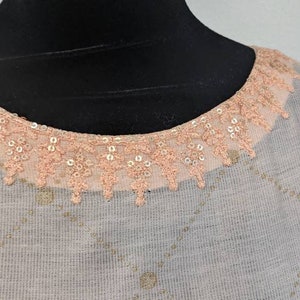 May include: A light-colored top with a round neckline trimmed with peach-colored lace and sequins. The fabric has a subtle diamond pattern with gold-colored accents. The top is displayed on a black mannequin.