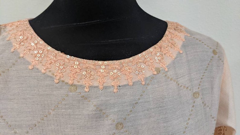 May include: A light-colored top with a round neckline trimmed with peach-colored lace and sequins. The fabric has a subtle diamond pattern with gold-colored accents. The top is displayed on a black mannequin.