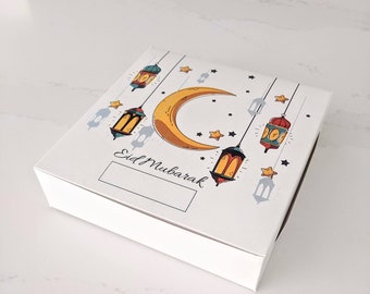 May include: A white square gift box with a decorative top featuring a crescent moon, stars, and hanging lanterns in shades of yellow, orange, green, and blue. The words "Eid Mubarak" are written on the box.